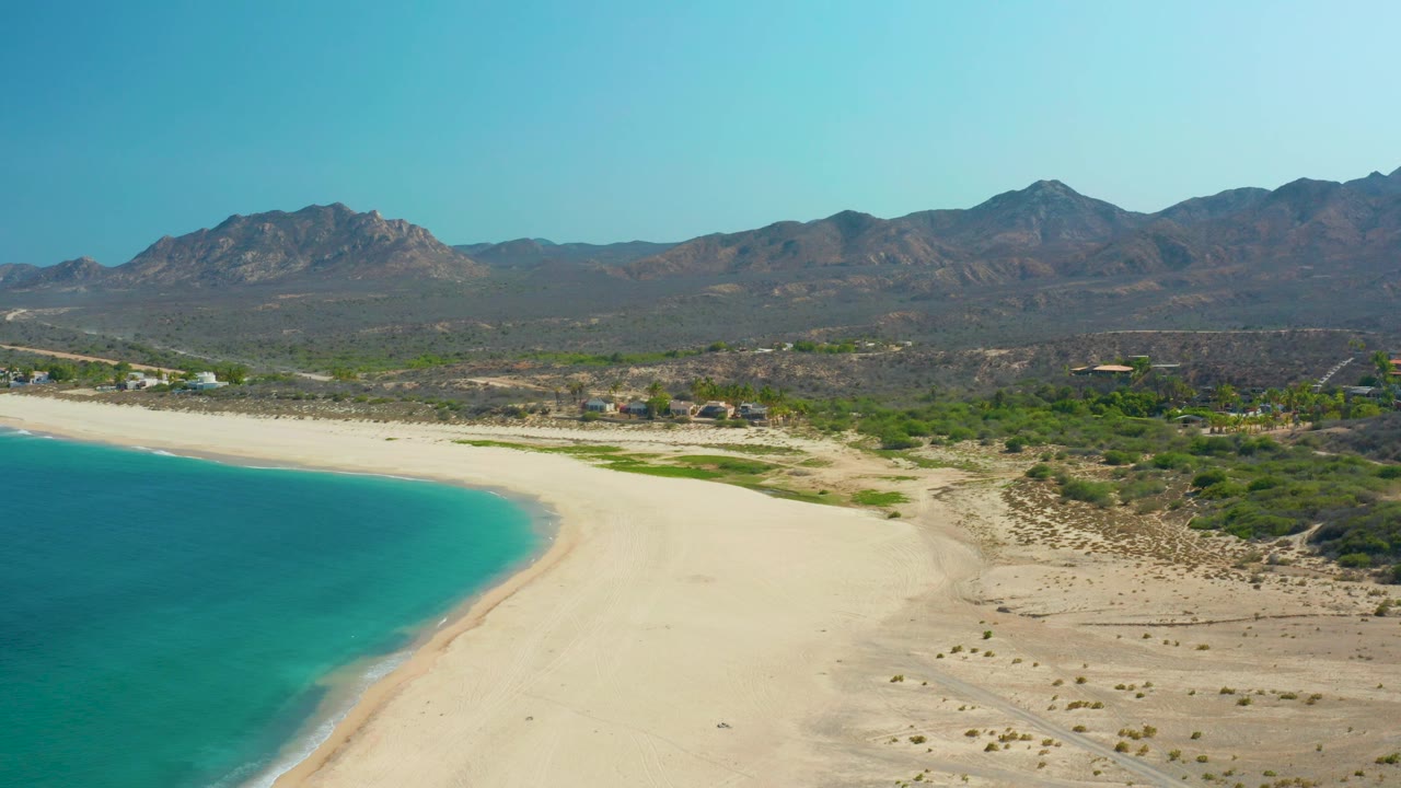 Opening frame of Los Frailes, Los Cabos, with turquoise water at left, pale sand across the center, desert scrub and distant mountains under clear sky, and no visible people.