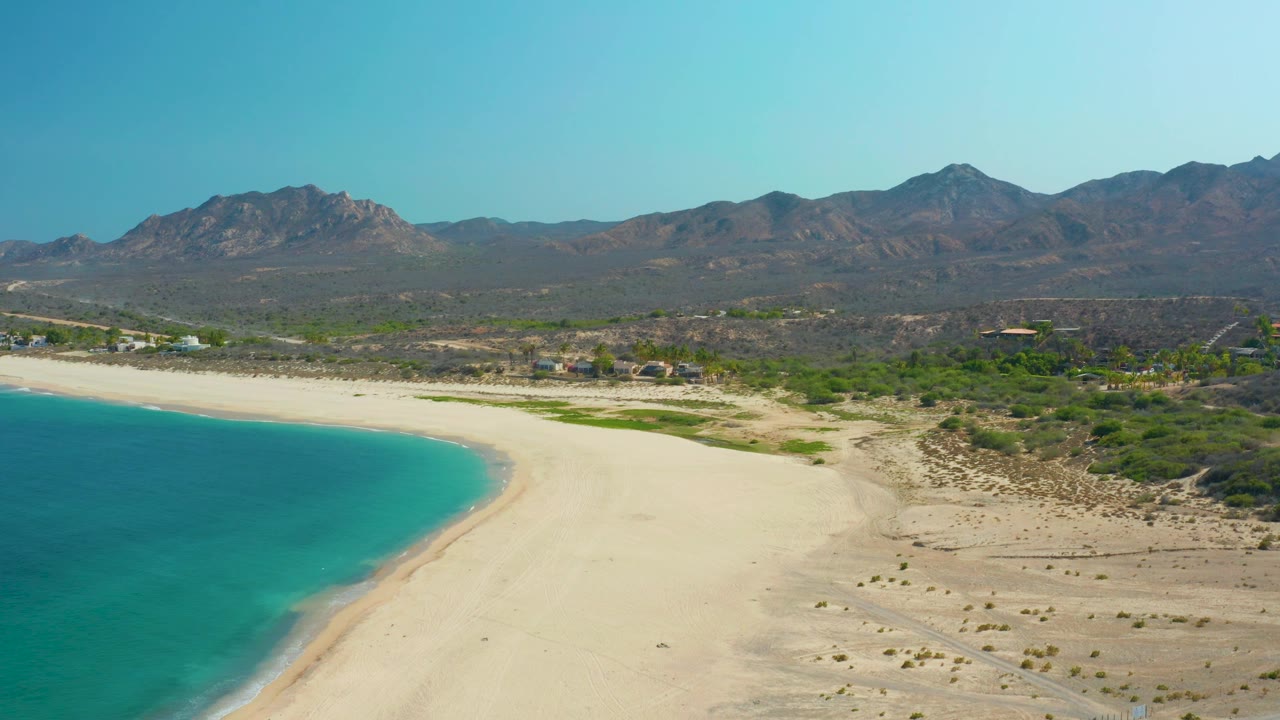 A broad Los Frailes beach view with the sea on the left, unoccupied sand in the center, green desert vegetation and a few distant structures inland, mountains behind, and no visible people.