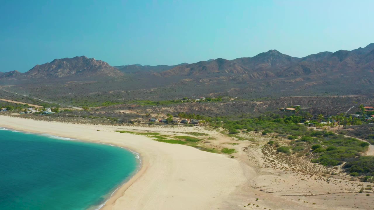 Los Frailes aerial frame showing blue-green water along the left edge, pale sand across the lower frame, brushy terrain, small buildings, mountains, and no visible people.