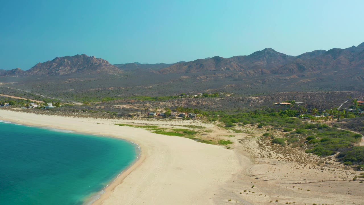 Los Frailes coastline seen from above with the ocean filling the lower left, a broad sand strip, patches of green scrub, small buildings, mountains, and no visible people.
