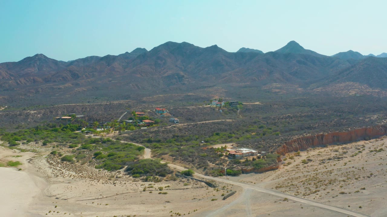 Aerial view over Los Frailes desert terrain with a pale dirt road, scattered shrubs, small buildings, rust-colored bluffs, distant mountains, and no visible people.