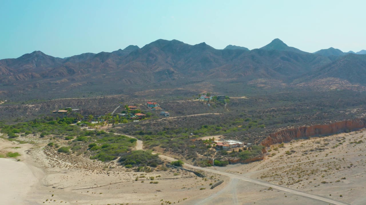 Wide aerial frame in Los Frailes showing the winding dirt road crossing sandy ground toward brush, scattered buildings, dark mountain ridges, and no visible people.