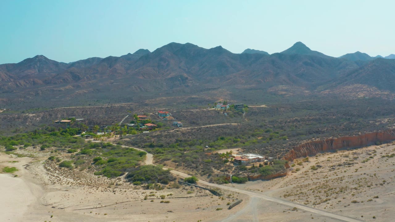 Aerial view of Los Frailes with sandy road tracks, pockets of green vegetation, scattered buildings, a reddish bluff line, distant mountains, and no visible people.