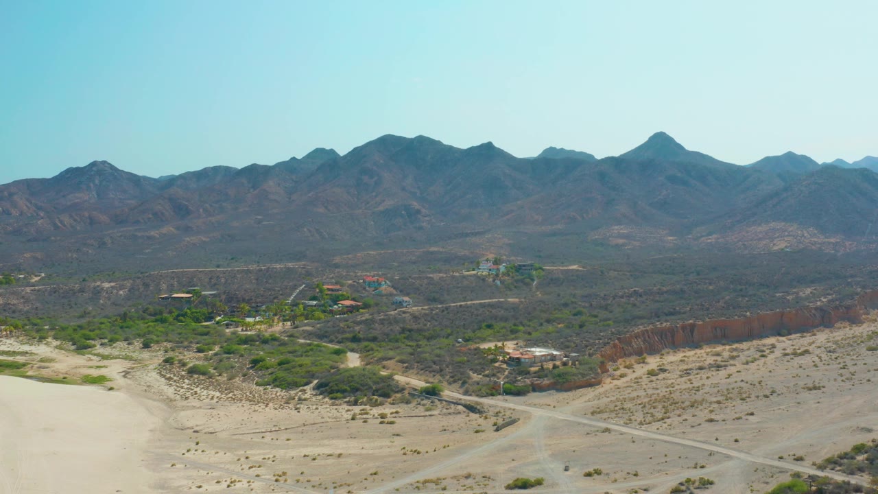Aerial view over Los Frailes desert sand, scrub, dirt roads, scattered buildings, and a dark mountain range under clear blue sky, with no visible people.