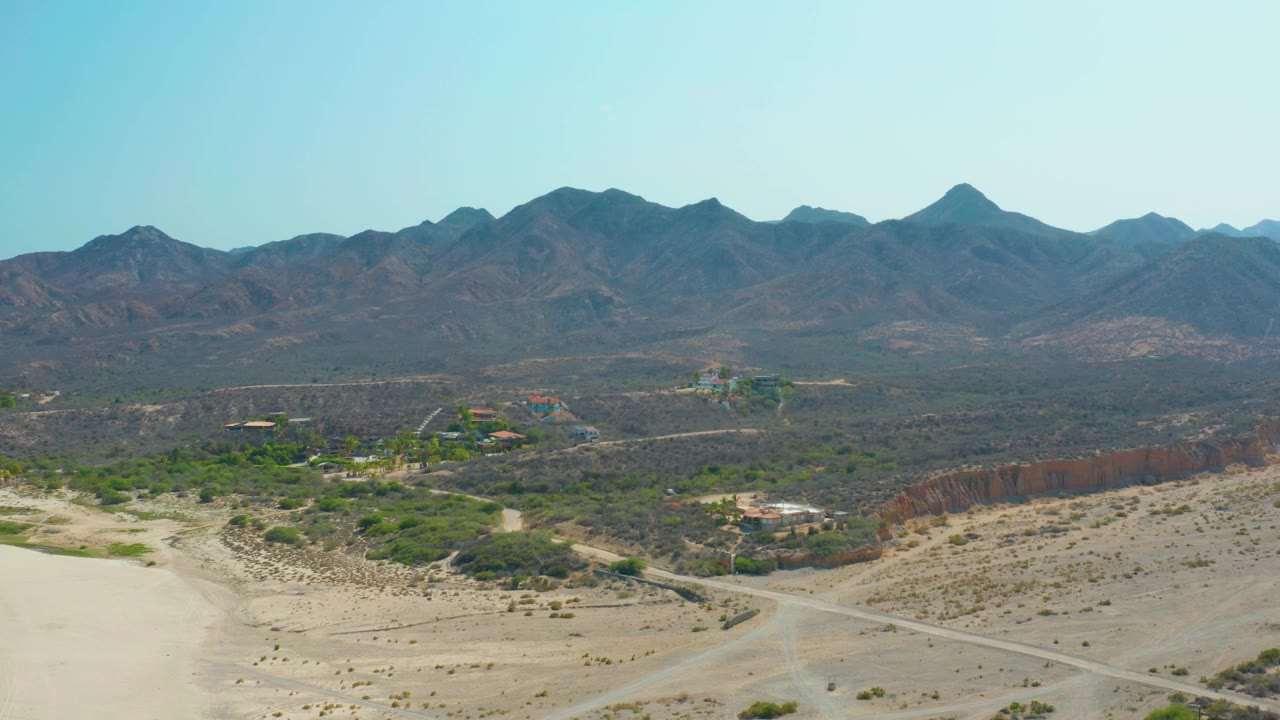 Wide aerial over Los Frailes showing sandy foreground terrain, a road crossing the frame, green scrub patches, scattered buildings, a red earth edge, and no visible people.