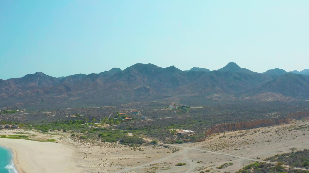 Opening frame over Los Frailes, Baja California Sur, with turquoise water and pale sand at left, scrubby desert roads across the foreground, mountains on the horizon, and no visible people.