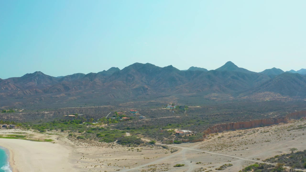Los Frailes aerial view with the beach nearly at the lower left edge, a winding dirt road crossing sandy desert, low green scrub in the midground, mountains beyond, and no visible people.
