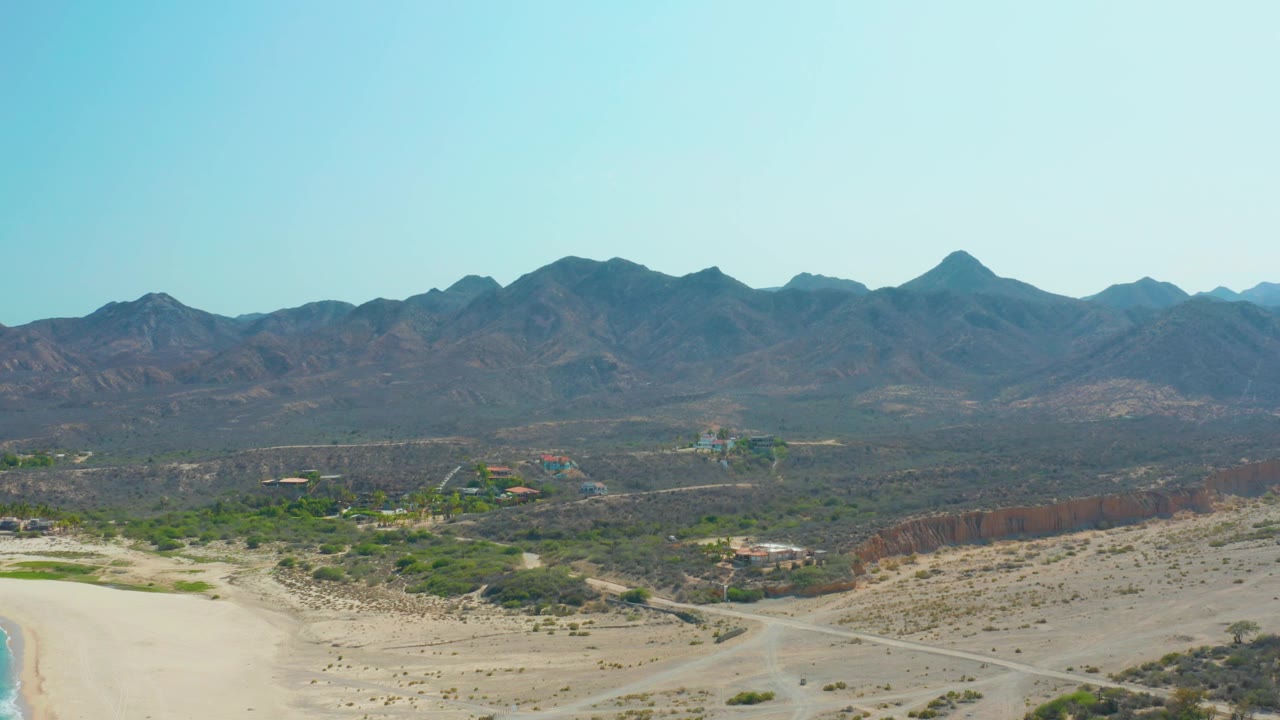 Drone view of arid bushland, dirt roads, and mountains with beach slipping out of frame.