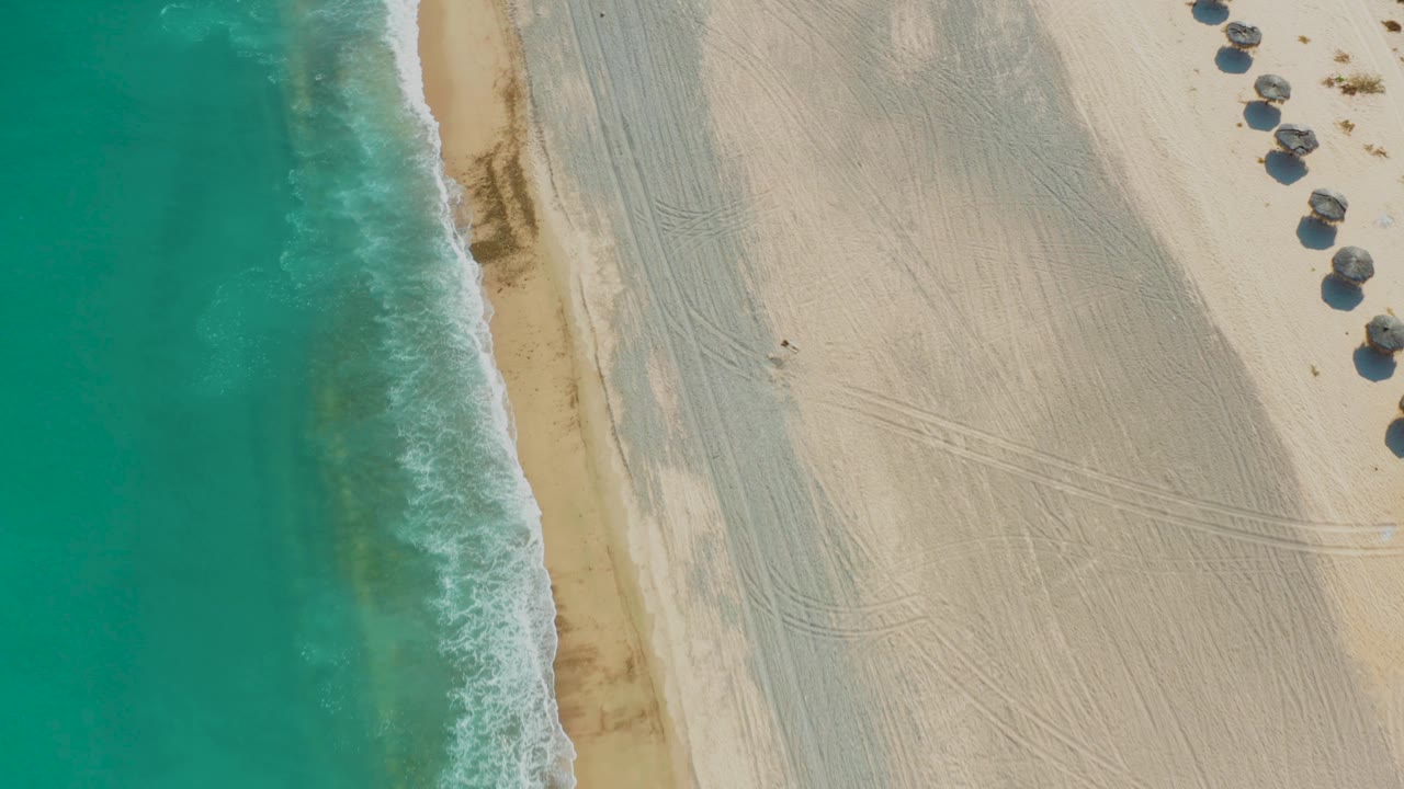 Aerial frame over Los Frailes showing turquoise water pushing a foamy edge onto darker wet sand, broad pale beach with track marks, huts at the upper right edge, and no visible people.
