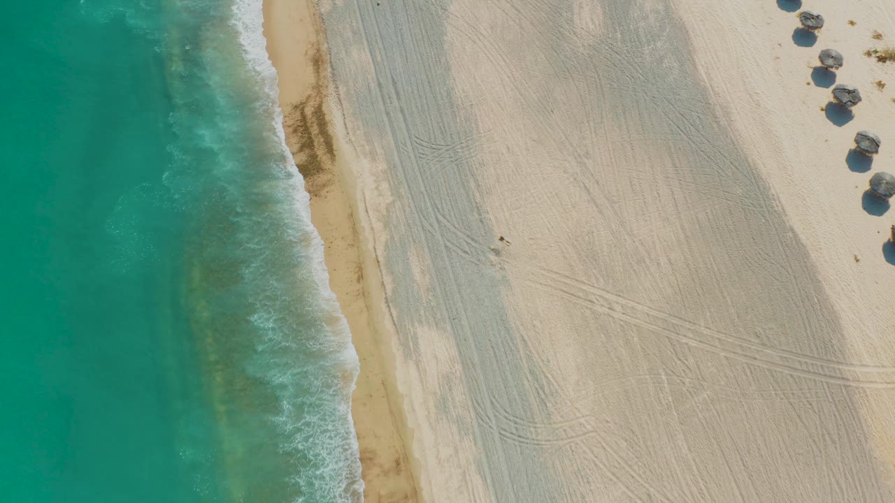 Top-down view of the Los Frailes shoreline as the foam line sits farther into the frame, with turquoise ocean left, pale raked sand right, huts near the upper right, and no visible people.