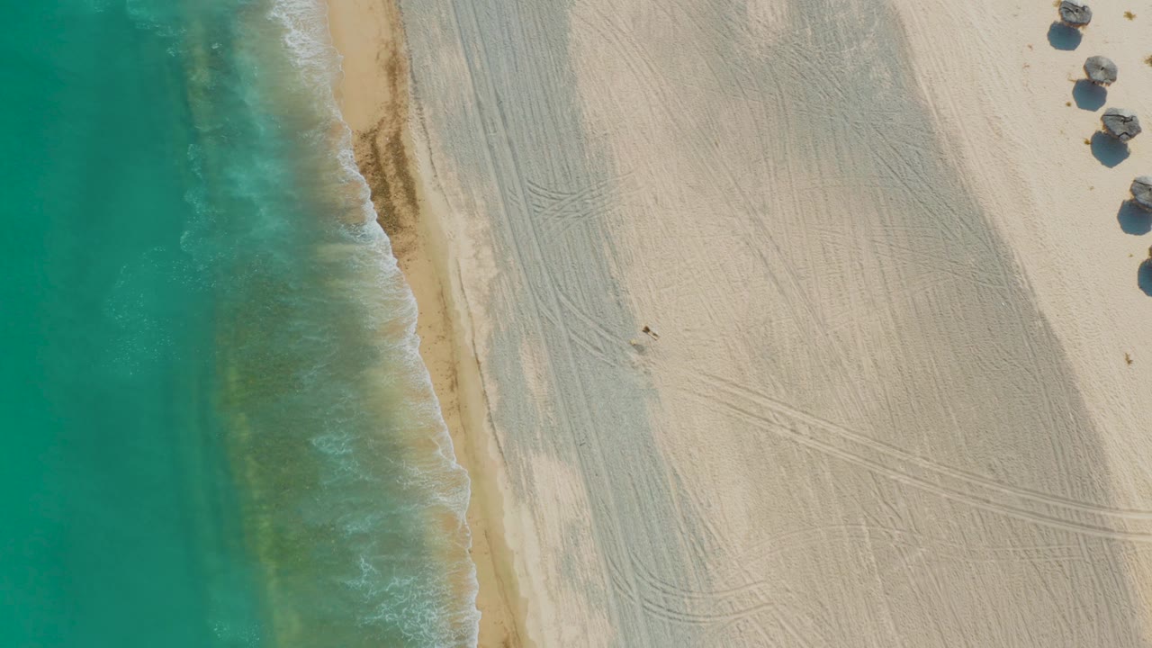 Overhead Los Frailes beach frame with more water visible on the left, a narrow band of foam along the shore, pale sand across the right, partial huts and shadows at the upper right, and no visible people.