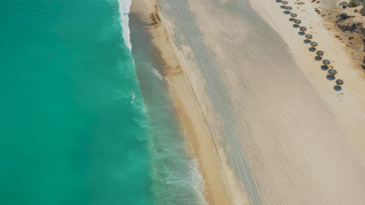 Aerial view over Los Frailes beach in Baja California Sur, with turquoise water on the left, pale sand on the right, a thin surf line near the shore, and no visible people.