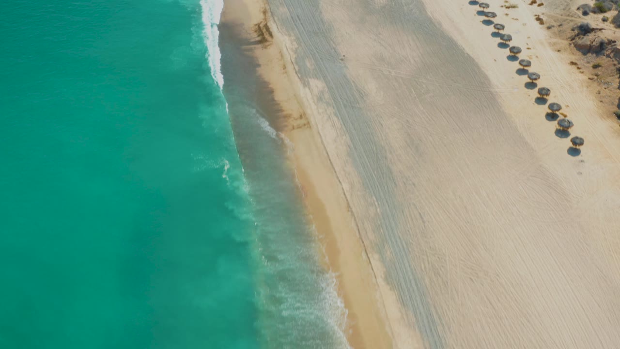 Overhead coastline with turquoise ocean, pale beach sand, and a row of umbrellas near the dunes.