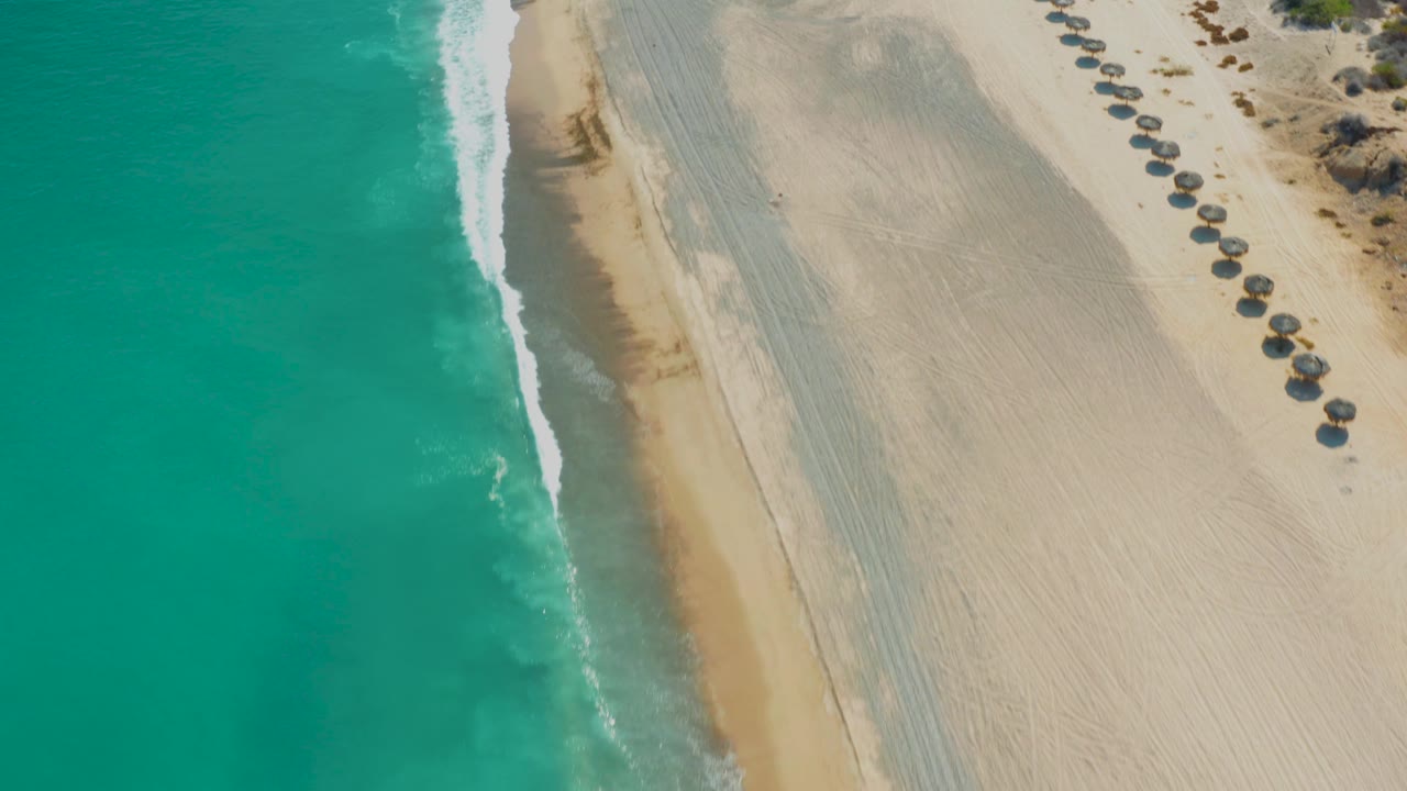 High aerial frame of Los Frailes beach, showing green-blue water, a slanted strip of wet sand, textured dry sand, umbrella shapes near the upper right edge, and no visible people.