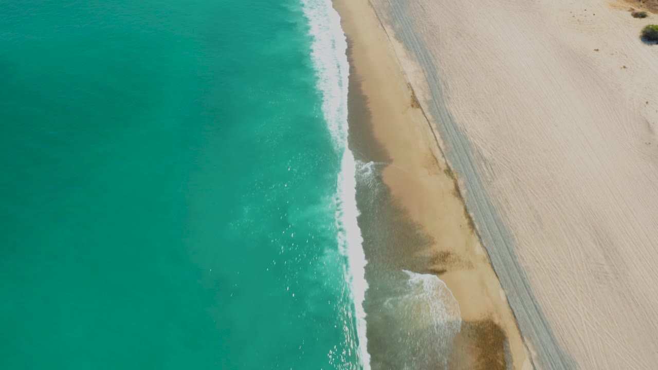 Top-down Los Frailes shoreline view with broad turquoise water, pale sand, a parallel track near the surf, and no visible people.