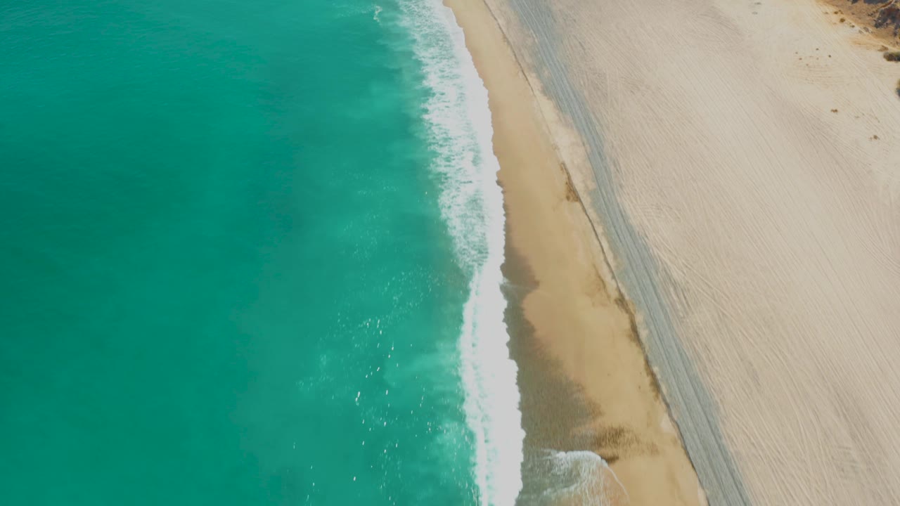 Overhead frame at Los Frailes where white foam spreads along the shoreline beside a clear strip of sand and a visible beach track, with no visible people.