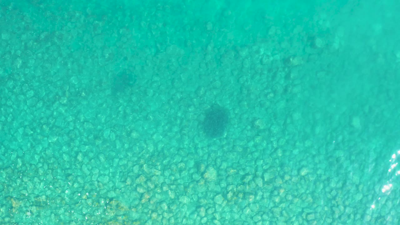 Clear turquoise ocean water reveals the rocky bottom while bright surface glints appear near the lower right, with no visible people.