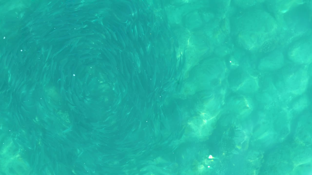 One second in, the overhead view shows turquoise shallow water, a softened circular swirl, and refracted light over submerged rocks in Los Frailes, with no visible people.