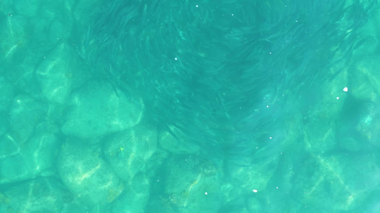 A top-down view of turquoise water reveals rounded rocks below and early swirl patterns on the surface, with no visible people.