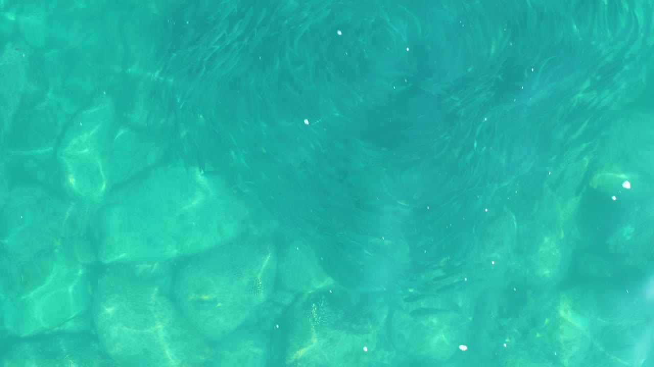 Swirling darker ripple shapes spread across shallow turquoise water over pale rocks in Los Frailes, with no visible people.