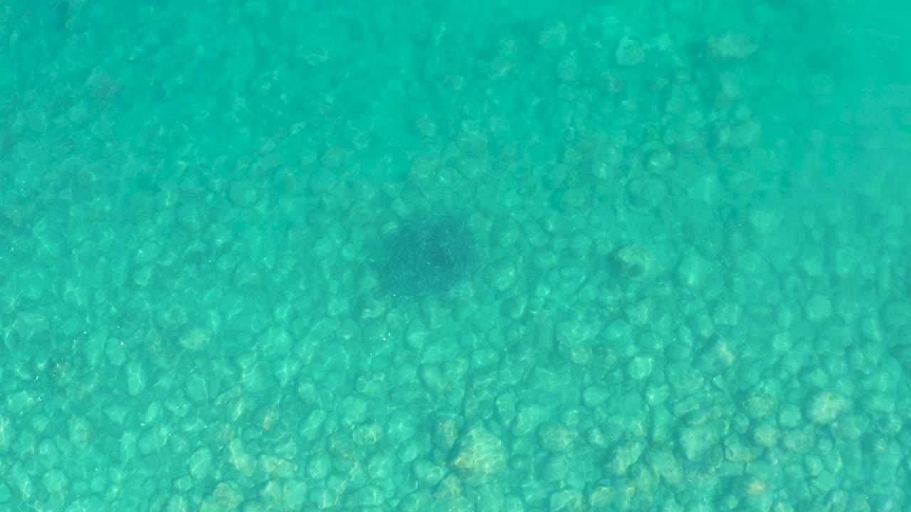 A darker soft-edged shadow patch appears over the turquoise water and stony seabed in Los Frailes, Los Cabos, with no visible people.
