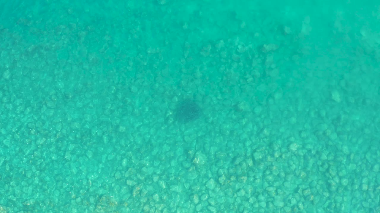 Turquoise water covers the rocky seabed in Los Frailes, Baja California Sur, with no visible people and a faint darker shape beginning to read against the stones.