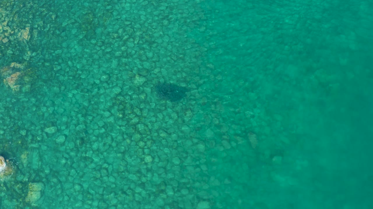 Turquoise water over a dense field of rocks in Los Frailes, with a dark underwater patch right of center and no visible people.