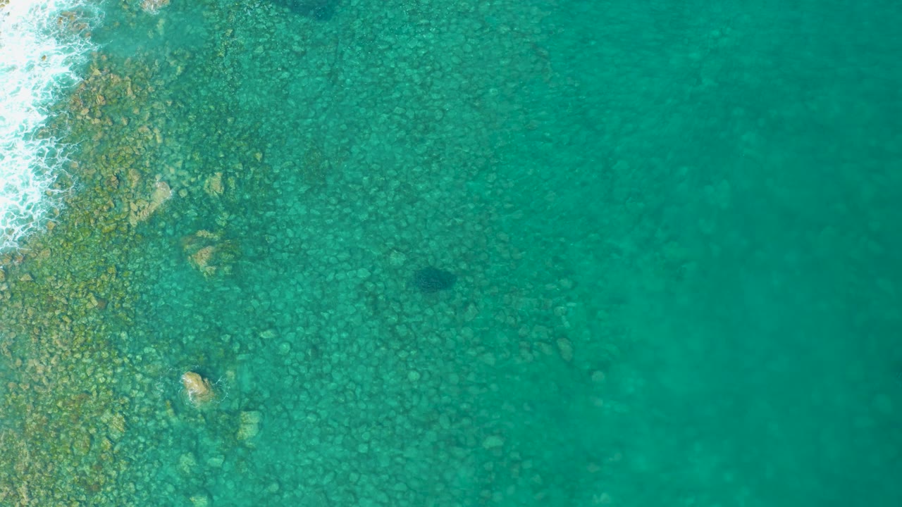 Second overhead frame in Los Frailes showing clustered submerged rocks under clear green-blue water, with a narrow strip of white foam on the left and no visible people.