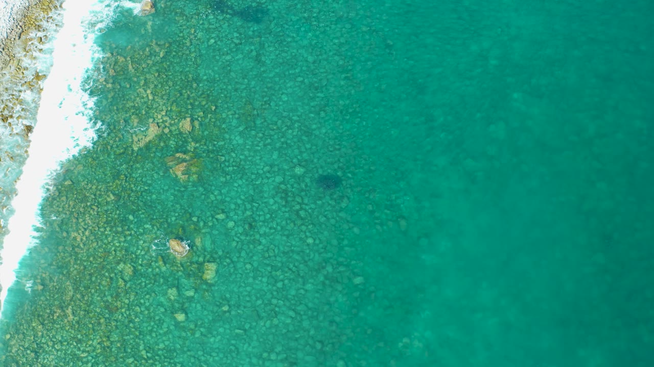 Straight-down aerial view in Los Frailes showing a bright white wave line along the left border, rocky shallows beneath turquoise water, and no visible people.