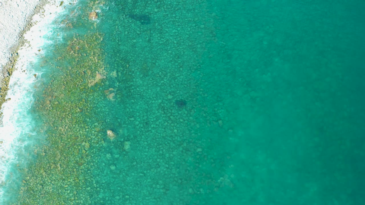Overhead view of Los Frailes turquoise coastal water with pale sand and rocky shallows along the left edge, no visible people.