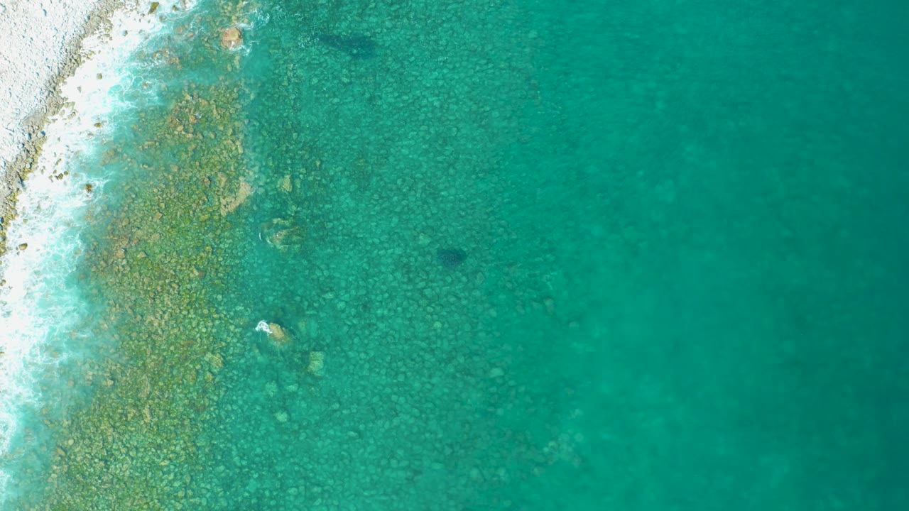 Overhead Los Frailes coastal water with a rocky shelf near the left shoreline and smoother turquoise water across the right side, no visible people.