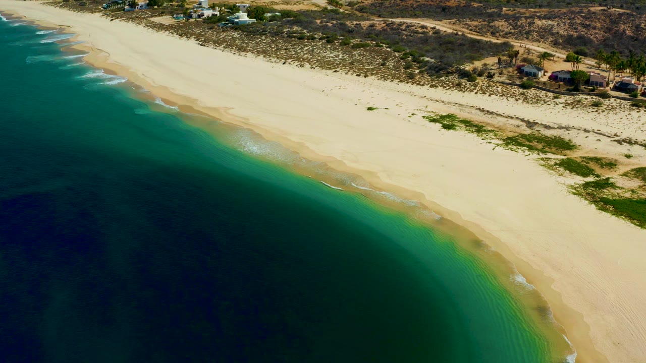 Aerial frame over Los Frailes, Los Cabos showing dark blue water, green shallows, pale sand, scrub vegetation, and small structures near the top edge, with no visible people.