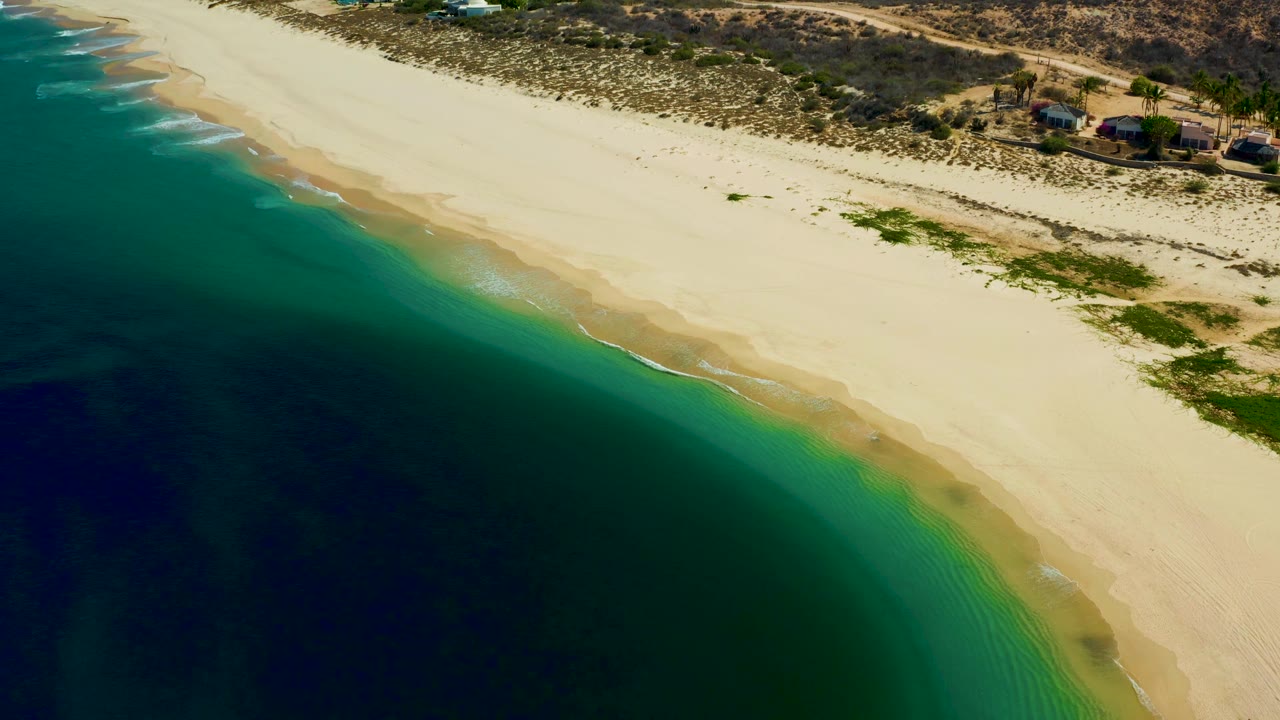 Aerial frame of the Los Frailes shoreline with a pale beach running diagonally, deep water at lower left, green shallows near shore, scrubland, and small buildings at the top, with no visible people.