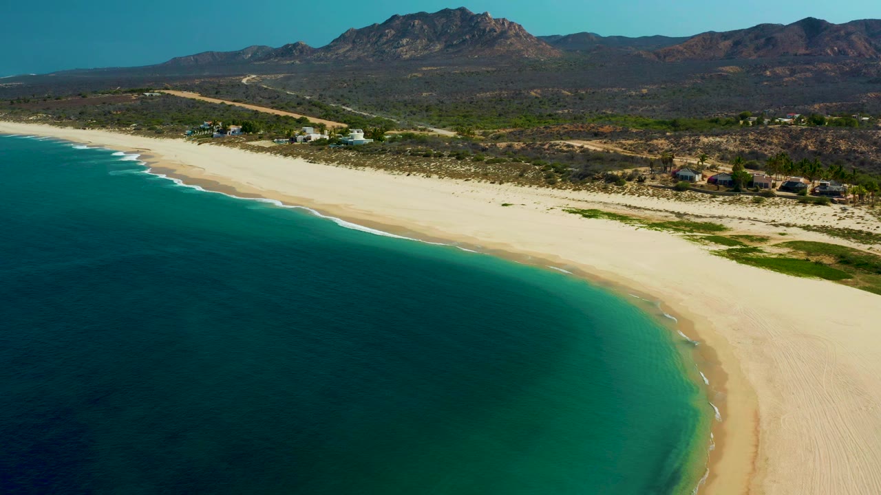 Opening aerial frame over Los Frailes, Baja California Sur, with turquoise ocean at left, pale sand curving across the shore, mountains in the background, and no visible people.