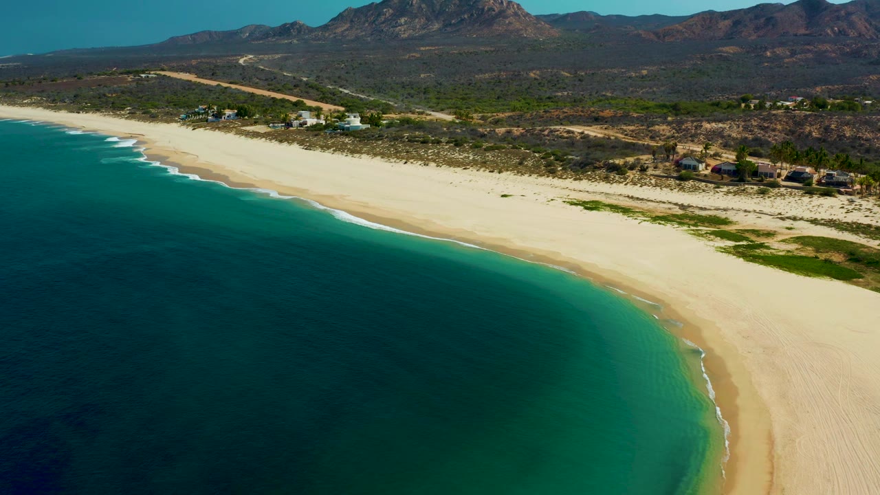 Aerial frame in Los Frailes showing the shoreline curve more prominently, with dark blue water offshore, bright sand along the beach, scrubland behind it, and no visible people.