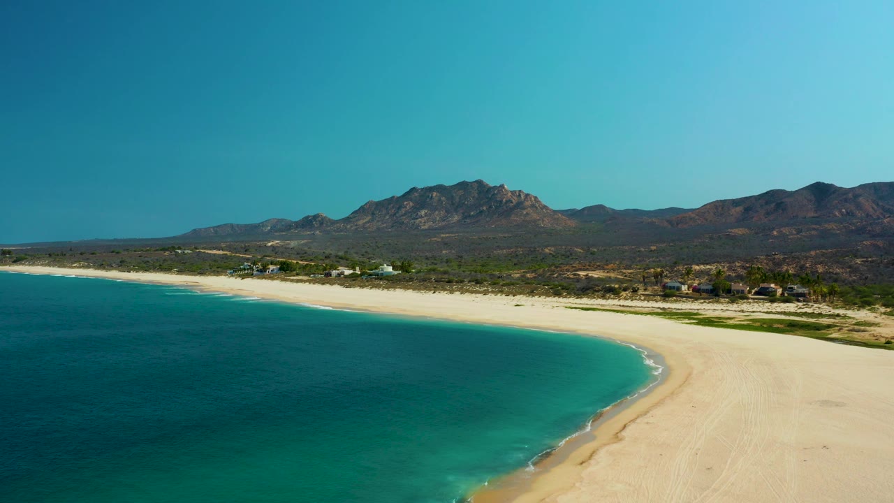 Aerial frame over Los Frailes showing the turquoise bay on the left, a wide pale beach sweeping to the right, low surf at the waterline, distant mountains, and no visible people.