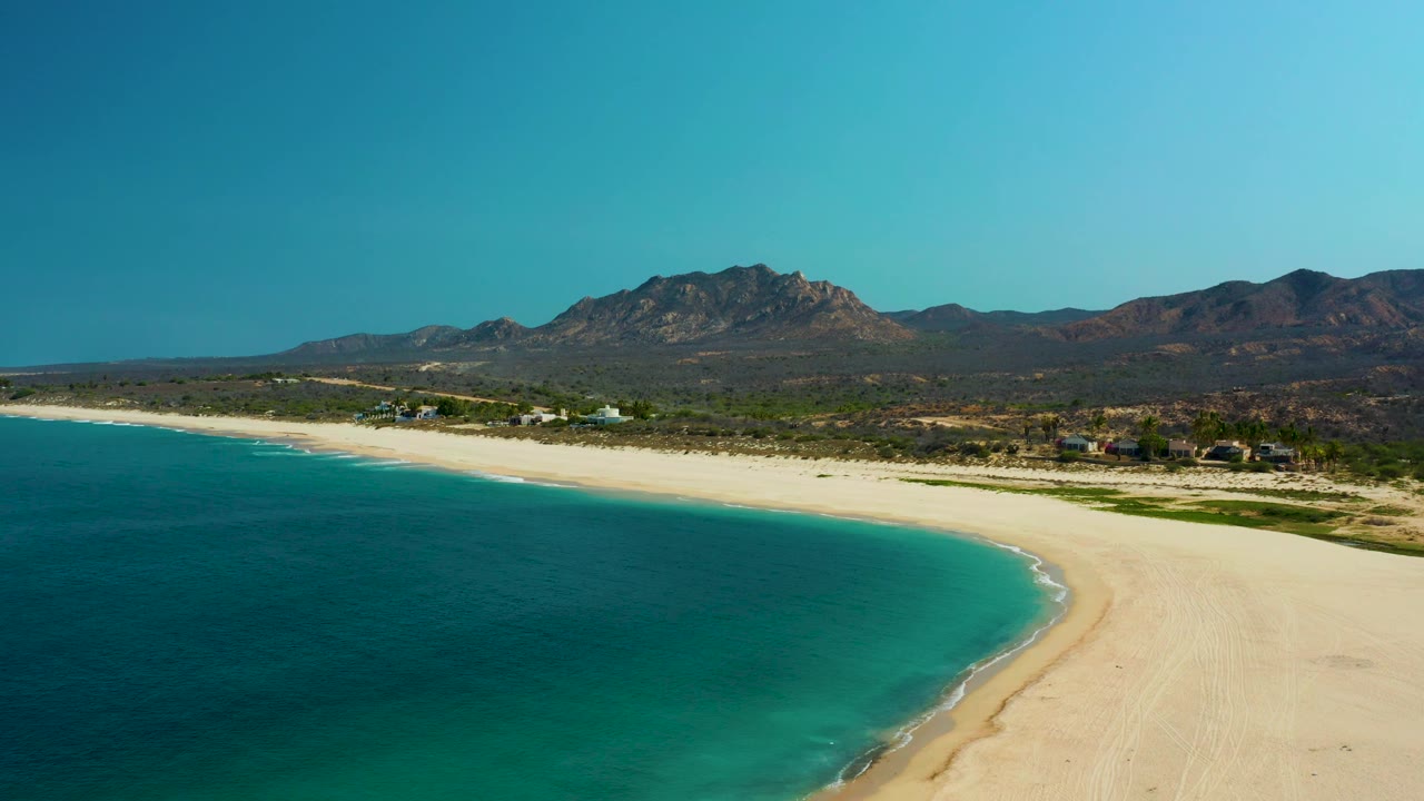 Drone frame over Los Frailes with teal water in the lower left, bright sand along the coast, scattered small buildings near the dunes, mountains behind them, and no visible people.