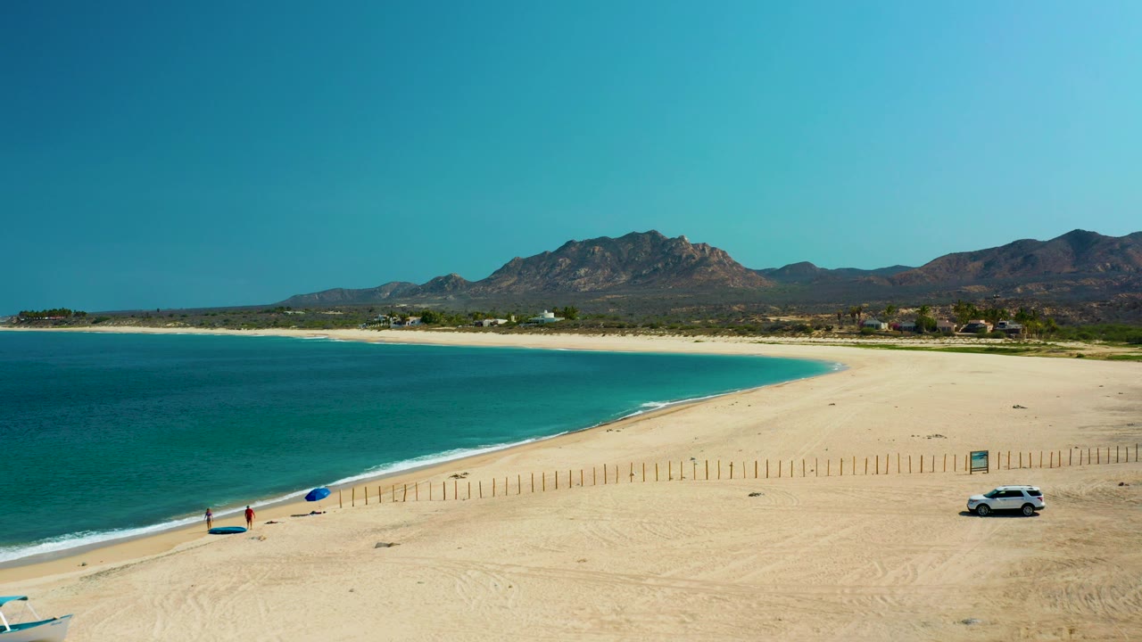 Aerial view of Los Frailes beach with turquoise water at left, pale sand, a blue umbrella, tiny beachgoers, a white car near fence posts, and mountains under clear sky.