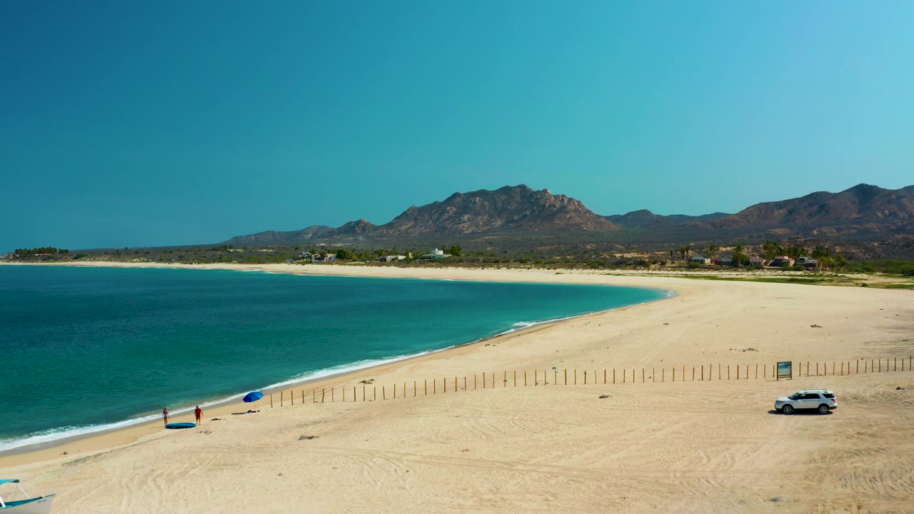 Curved sandy beach with turquoise bay, umbrella, white car, and mountains.