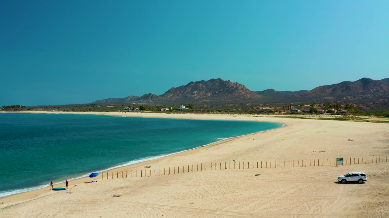 Aerial frame over Los Frailes beach with turquoise water filling the left side, fence posts crossing the sand, a white car near the right edge, tiny people near the shoreline, and mountains in back.