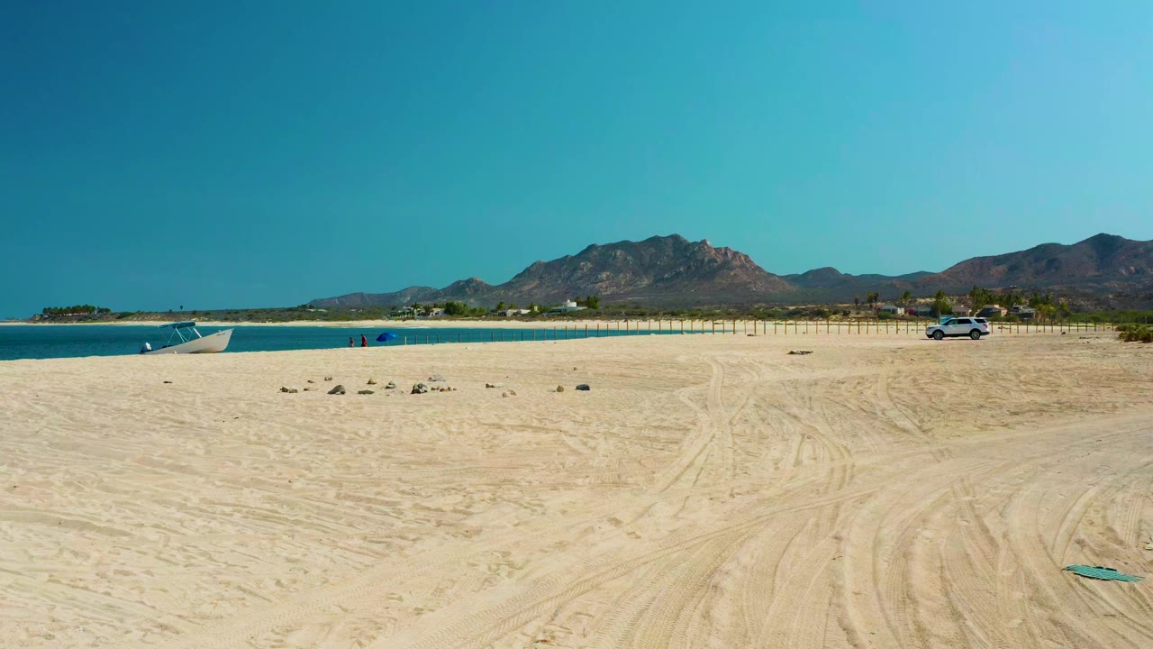 Wide aerial view of Los Frailes beach with tire-marked sand in front, turquoise water on the left, distant mountains, a small boat near shore, a parked car to the right, and no visible people.