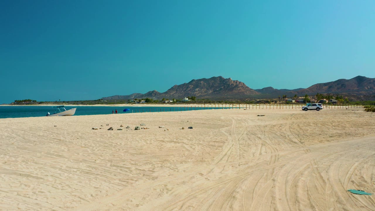 Los Frailes beach from above with broad sand in the foreground, ocean and a small boat on the left edge, a parked car near the right fence line, distant mountains, and no visible people.