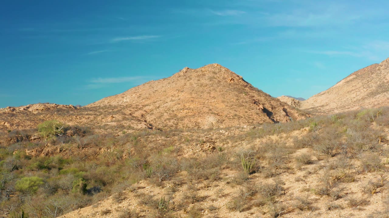 Mid-clip view of Cabo Pulmo arid terrain with sandy foreground ridges, scattered bushes, rocky mounds, and open blue sky, with no visible people.