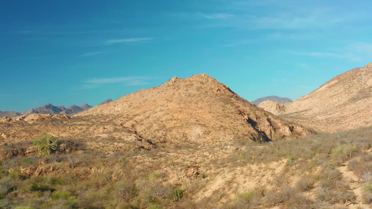 Bright Cabo Pulmo desert landscape with a broad rocky hill, low scrub, cactus-like plants, distant left ridges, and no visible people.