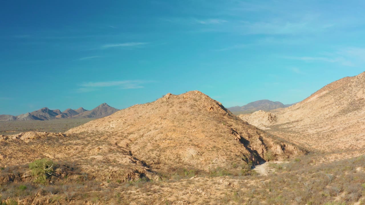 Start of a Cabo Pulmo desert landscape with sandy scrub, a central tan rocky hill, distant mountains at left, clear blue sky, and no visible people.