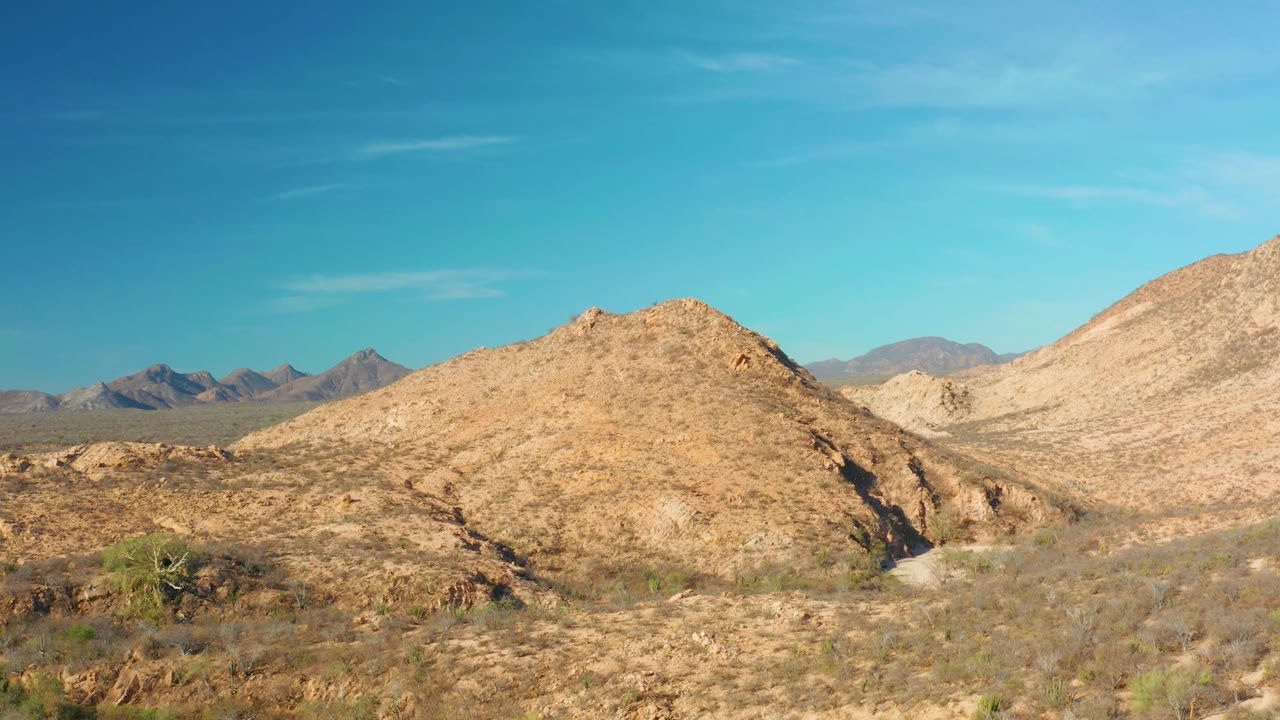 Early wide frame from Cabo Pulmo with sparse bushes across sandy ground, a rounded rocky hill in the center, pale slope at right, distant mountains at left, and no visible people.