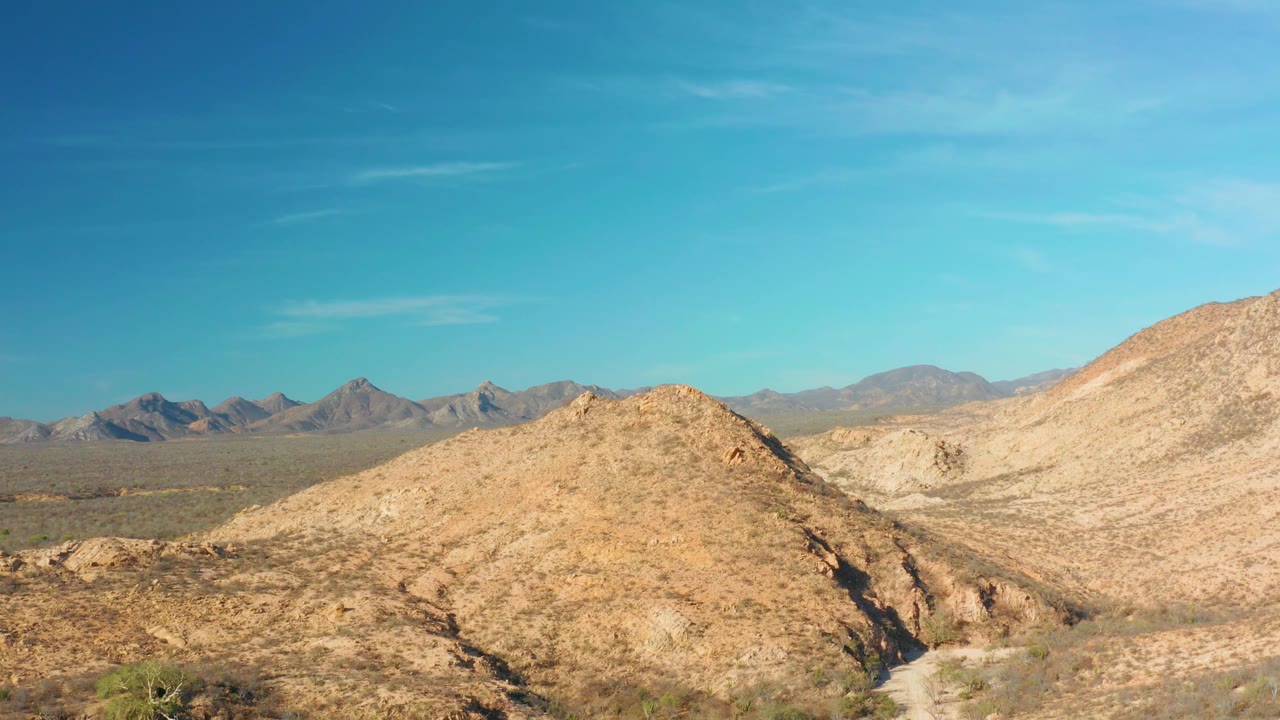Dry rocky terrain near Cabo Pulmo, Baja California Sur, shows a central tan hill, scattered shrubs at the lower edge, distant ridges, and no visible people.
