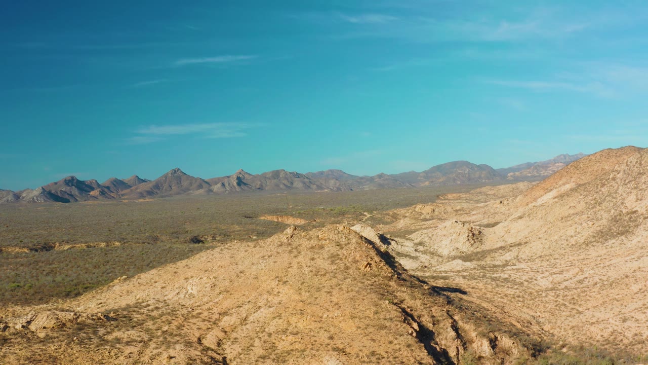 Cabo Pulmo desert terrain fills the foreground with a rocky slope to the right, low scrub across the plain, distant mountains, and no visible people.