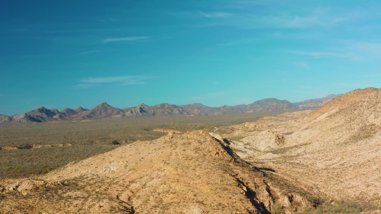 Cabo Pulmo desert ridges sit below a wide blue sky, with a darker rocky channel cutting through the foreground and distant mountains beyond, no visible people.
