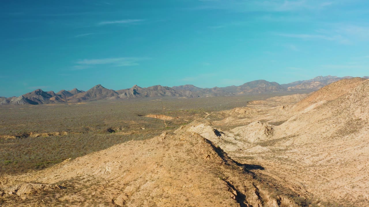 Cabo Pulmo aerial frame showing tan hillside terrain on the right, a dark wash cutting through the foreground, distant mountains, clear blue sky, and no visible people.
